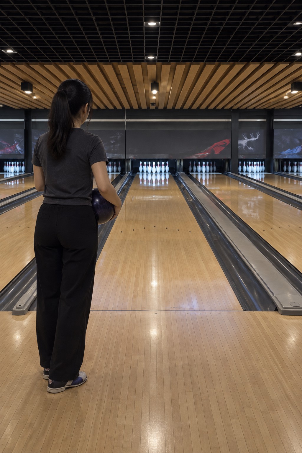 Portrait view straight along the central playing lane. Its full width leads from the foul line to a clear white pin rack; an adult woman stands on the left of the same approach.