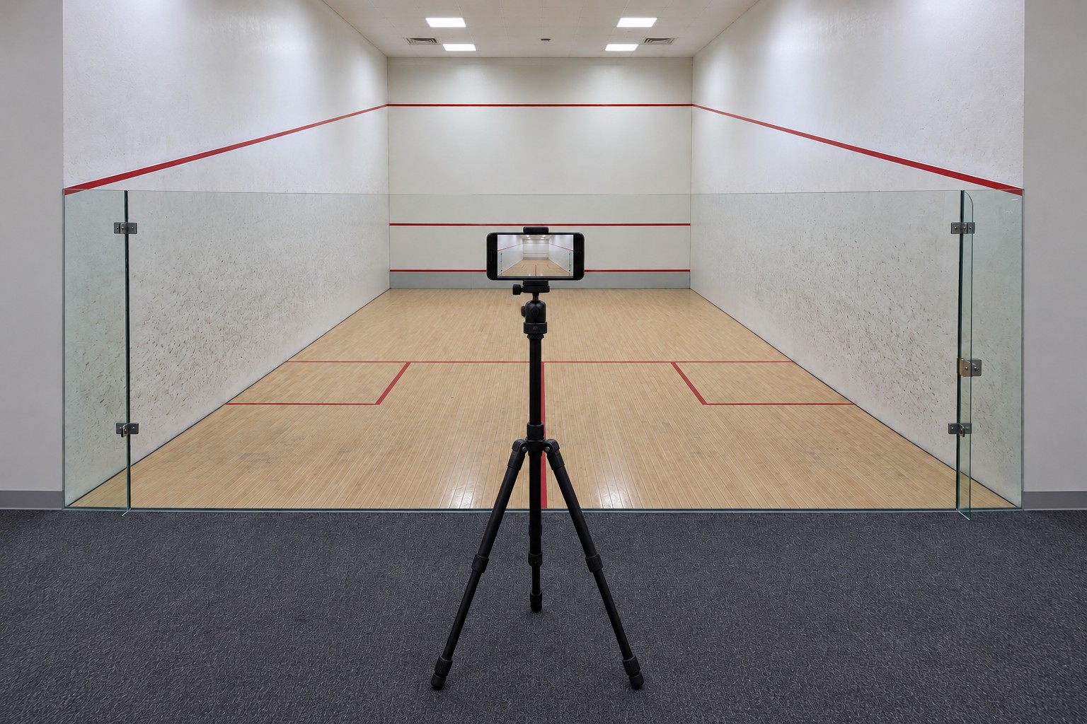 Full tripod on the spectator side of a squash court’s rear glass, with a horizontal phone aimed into the court and all three feet outside the playing area.